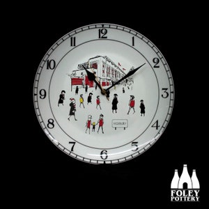 Puede incluir: Reloj de cerámica blanca con una esfera negra y agujas negras. La esfera del reloj presenta una ilustración en blanco y negro de un estadio con personas caminando hacia él. La esfera del reloj también incluye los números 12, 1, 2, 3, 4, 5, 6, 7, 8, 9, 10 y 11. La esfera del reloj también incluye el texto "HICHBURY".