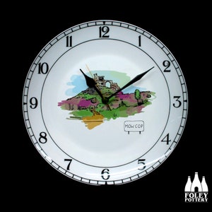 May include: White ceramic clock plate with black numbers and hands. The clock face features a watercolor illustration of a castle on a hill with the words "MOW COP" on a sign. The clock is made by Foley Pottery.