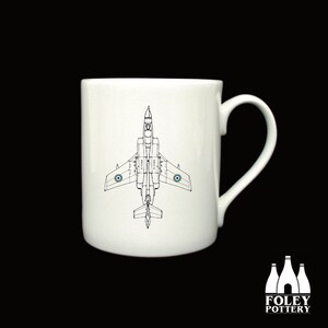 Può includere: Una tazza in ceramica bianca con un disegno a linea nera di un aereo a reazione. L'aereo è rivolto verso il basso con le ali spiegate. La tazza ha un manico sul lato destro.