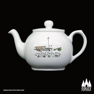 May include: White ceramic teapot with a black and white illustration of a baseball stadium with people in the stands. The teapot has a round body and a spout on the left side.