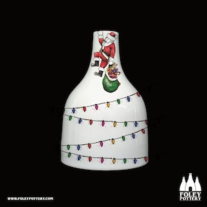 Pode incluir: Vaso de cerâmica branco com tema natalino. Papai Noel está escalando o vaso com uma sacola de presentes. O vaso é decorado com luzes de corda coloridas.