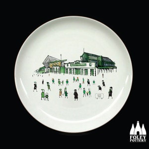 Puede incluir: Un plato de cerámica blanco con una ilustración verde y negra de un estadio y personas caminando hacia él. El texto "Hope Park" es visible en el plato.
