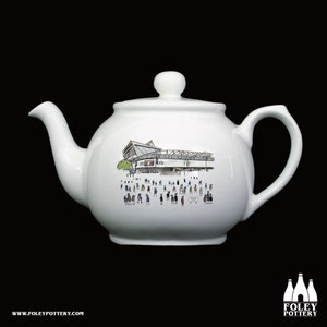 May include: White ceramic teapot featuring a black and white illustration of a stadium and people on the side. The teapot has a handle and a lid.