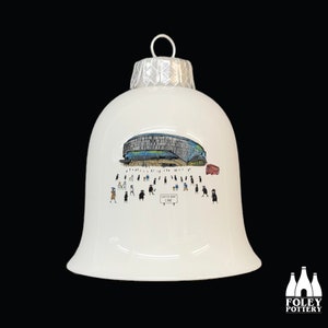 Puede incluir: Adorno navideño de cerámica blanca con forma de campana con una ilustración azul y gris de un edificio y personas. El texto "White Hart Lane" es visible en un cartel en la ilustración.