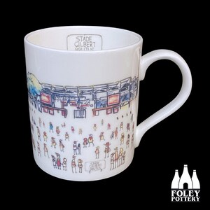 May include: White ceramic mug with a black and white illustration of a stadium and people. The text "Stade Gilbert Brutus" is printed on the mug.