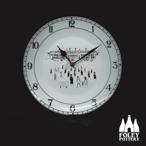 May include: A white ceramic clock with black numbers and hands. The clock face features a black and white illustration of Wembley Stadium with people walking towards it. The clock has the words "Wembley Stadium" printed on it. The clock is made by Foley Pottery.