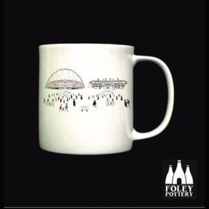 May include: White ceramic mug with a black and white illustration of a stadium and people. The mug has a black handle and the words "FOLEY POTTERY" are printed on the bottom.