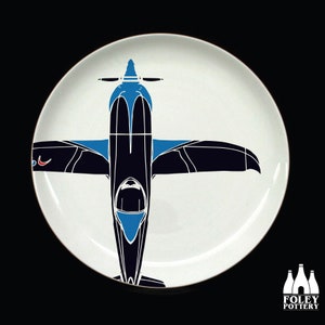 May include: A white ceramic plate with a black and blue silhouette of a plane. The plane is facing upwards with the wings spread out. The plane has a blue cockpit and a red stripe on the wing.