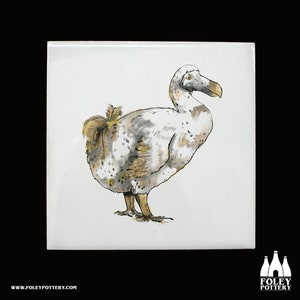 May include: A white ceramic tile with a hand-drawn illustration of a dodo bird in black, gray, and gold. The dodo is facing the right side of the tile and has a long beak.