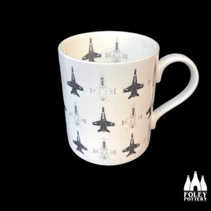 May include: A white ceramic mug with a black and white pattern of fighter jets. The mug has a handle on the right side.