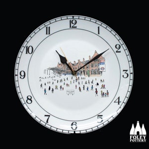 Puede incluir: Un plato de cerámica blanco con una esfera de reloj negra y agujas negras. El plato presenta una ilustración en blanco y negro de una estación de tren con gente caminando. El texto "MARYLEBONE STATION" es visible en el plato.