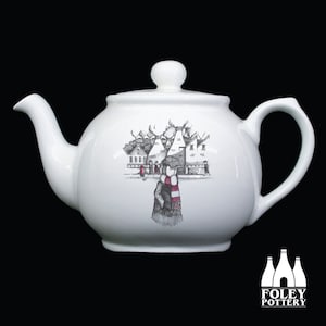 Può includere: Teiera in ceramica bianca con un'illustrazione in bianco e nero di una persona con una sciarpa rosso e bianca in piedi di fronte a una fila di edifici. Il testo "FOLEY POTTERY" è stampato sul fondo della teiera.