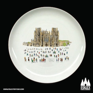 Puede incluir: Un plato de cerámica blanco con una ilustración dibujada a mano de la Catedral de Wells en Inglaterra. La catedral está representada en tonos marrones, amarillos y grises. Hay pequeñas figuras de palo de colores delante de la catedral. El texto "Wells Cathedral" está escrito en negro sobre un cartel blanco.