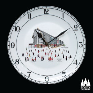 May include: White ceramic wall clock with black numerals and hands. The clock face features a detailed illustration of a building with people and the text "Brisbane Road". The clock has a black border and the Foley Pottery logo.