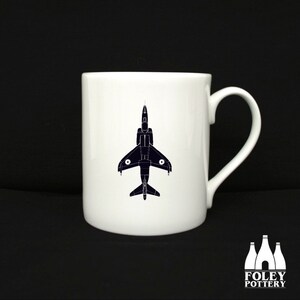 May include: White ceramic mug with a black silhouette of a jet plane on the front. The mug has a handle on the right side. The text "FOLEY POTTERY" is printed in white on the bottom of the mug.