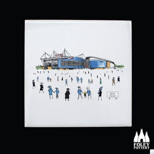 May include: A white ceramic tile with a hand-drawn illustration of a stadium with a blue roof and a sign that reads "Ricoh Arena".  There are many small stick figures in blue and white walking towards the stadium.