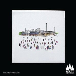 Peut inclure: Un carreau de céramique blanc avec une illustration en noir et blanc d'un stade avec une foule de personnes devant. Le texte "Don't Be So Trolley" est sur une pancarte tenue par l'une des personnes de la foule.
