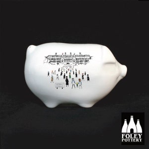 May include: White ceramic piggy bank with a black and white illustration of Wembley Stadium and a crowd of people walking towards it. The sign reads "Wembley Stadium".