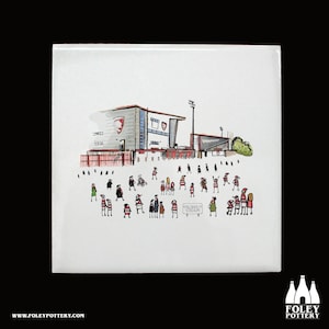 Puede incluir: Un azulejo de cerámica blanco con una ilustración dibujada a mano de un estadio con una multitud de personas con camisas a rayas rojas y blancas. El estadio está etiquetado como "Kincsholm Stadium".