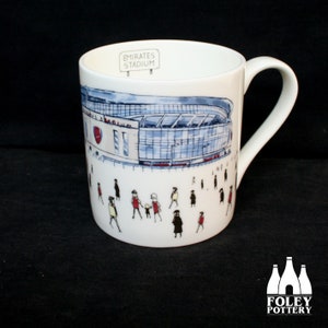 Puede incluir: Taza de cerámica blanca con un dibujo del Emirates Stadium, hogar del Arsenal Football Club. El estadio está representado en azul y gris con un escudo del Arsenal rojo y blanco. La taza también tiene un cartel que dice "Emirates Stadium".