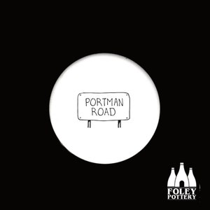 May include: A white ceramic coaster with a hand-drawn illustration of a road sign that reads "Portman Road" in black. The coaster has a black background and the Foley Pottery logo in the bottom right corner.