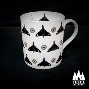 Puede incluir: Taza de cerámica blanca con un patrón en blanco y negro de aviones y dianas rojas y blancas. La taza tiene un asa blanca. La taza está hecha por Foley Pottery.