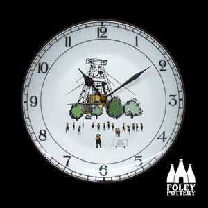 May include: A white ceramic wall clock with a black rim, featuring a hand-drawn illustration of a mining structure, trees, and figures. The clock has black hands and numerals. The text "HEM HEATH" is visible.