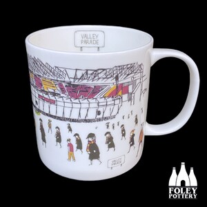 Puede incluir: Taza de cerámica blanca con una ilustración de estadio y el texto "Valley Parade". La taza tiene un asa y una ilustración en blanco y negro de personas caminando. El logotipo de Foley Pottery está en la parte inferior.
