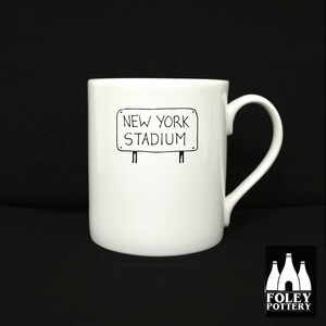 May include: White ceramic mug with a black hand-drawn illustration of a sign that reads "NEW YORK STADIUM".