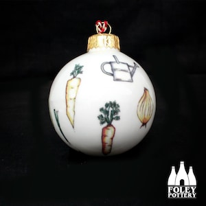 May include: White ceramic Christmas ornament with a gold top and a red and white string. The ornament is decorated with a variety of vegetables, including carrots, onions, and parsley.