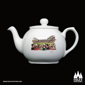 Puede incluir: Tetera de cerámica blanca con una ilustración de dibujos animados de una escena de estadio con aficionados con camisetas rojas y blancas. El texto "Anfield" es visible en la tetera.