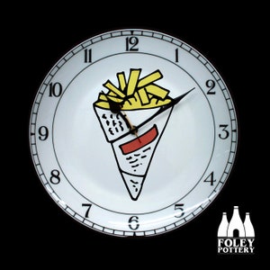 May include: White ceramic clock with black numbers and hands. The clock face features a black and yellow illustration of a cone of french fries. The clock is made by Foley Pottery.