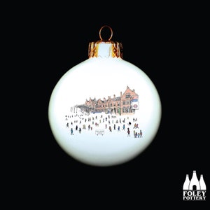 Puede incluir: Adorno navideño de cerámica blanca con una ilustración dibujada a mano de una estación de tren con gente caminando. El adorno tiene una parte superior dorada.