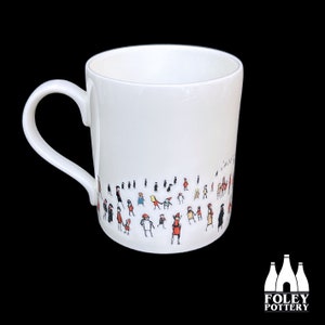 May include: White ceramic mug with a black handle. The mug has a black and white illustration of a crowd of people around the bottom edge.
