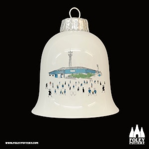 May include: White ceramic Christmas ornament shaped like a bell with a blue and white illustration of a stadium and people. The text "The City" is visible on the ornament.