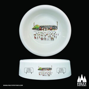 May include: White ceramic dog bowl with a black and white illustration of a stadium with people in the stands. The bowl has the text "Stade Gilbert Brutus" on a sign in the illustration.