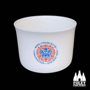 May include: White ceramic mug with a blue and red design commemorating King Charles III's coronation on May 6, 2023. The design features a crown with floral accents and the text "King Charles III Coronation 6th May 2023".