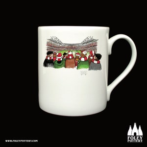 May include: White ceramic mug with a cartoon illustration of people wearing red and white scarves and hats watching a soccer game. The mug has a black handle and the text "SPURS" is visible on the bottom of the image.