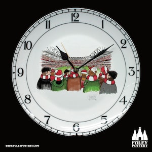 May include: A white ceramic clock with a black face and black numbers. The clock features a cartoon illustration of a football stadium with fans in the stands. The text "Emirates Stadium" is visible on a sign in the image.