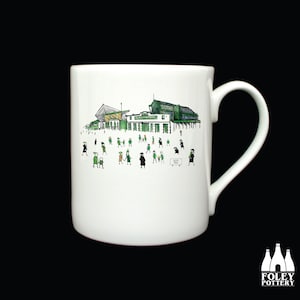 Puede incluir: Taza de cerámica blanca con una ilustración verde y negra de un estadio y personas. El texto "HOME PARK" es visible en un cartel.