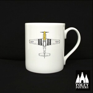 May include: White ceramic mug with a black and white illustration of a yellow and black airplane. The mug has a handle and the words "FOLEY POTTERY" are printed on the bottom.