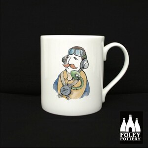 Könnte beinhalten: Weiße Keramiktasse mit einer Cartoon-Illustration eines Piloten in einem blauen Overall, einer Schutzbrille und einem Headset. Der Pilot hat einen Schnurrbart und schaut zur Seite. Die Tasse hat einen Henkel und die Worte "FOLEY POTTERY" sind auf dem Boden aufgedruckt.