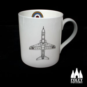 May include: White ceramic mug with a black line drawing of a jet plane on the front. The plane has a red, white, and blue roundel on the wings. The mug has a red, white, and blue roundel on the inside rim.