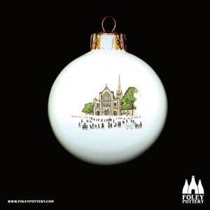 May include: White ceramic Christmas ornament with a hand-drawn illustration of a church building and people in front of it. The ornament has a gold top.