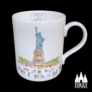 Può includere: Tazza in ceramica bianca con un'illustrazione colorata della Statua della Libertà e persone di fronte ad essa. Il testo "Statue of Liberty" è stampato sulla tazza.