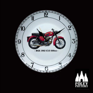 May include: A white ceramic clock with black numbers and a red and black motorcycle illustration. The clock face reads "BSA 1962 C15 500cc" and is made by Foley Pottery.