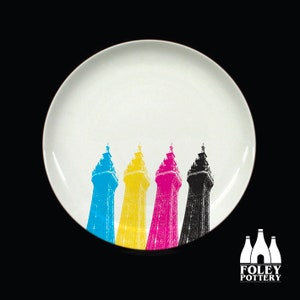 May include: A white ceramic plate with a black, white, yellow, and blue print of four towers. The towers are in a row and are printed in the colors of the CMYK printing process.