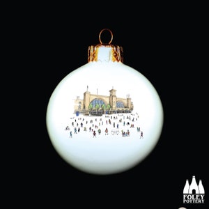Puede incluir: Adorno navideño de cerámica blanca con una ilustración dibujada a mano de la estación de King's Cross en Londres, Inglaterra. El adorno representa la fachada de la estación en detalle, con personas caminando alrededor de la estación.