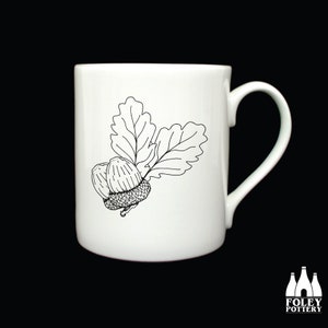 May include: White ceramic mug with a black line drawing of two acorns and an oak leaf.