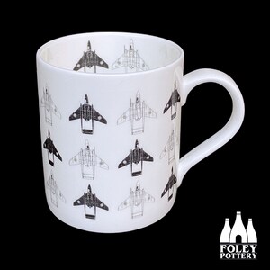 May include: A white ceramic mug with a black and white pattern of jet aircraft. The mug has a handle and the text "FOLEY POTTERY" is printed on the bottom.
